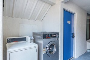 A laundry area with a white dryer, a front-loading washing machine, and a blue door marked with the number 122. - Motel 6 Cleveland, TX | 2 Queen Beds NS (Cleveland)
