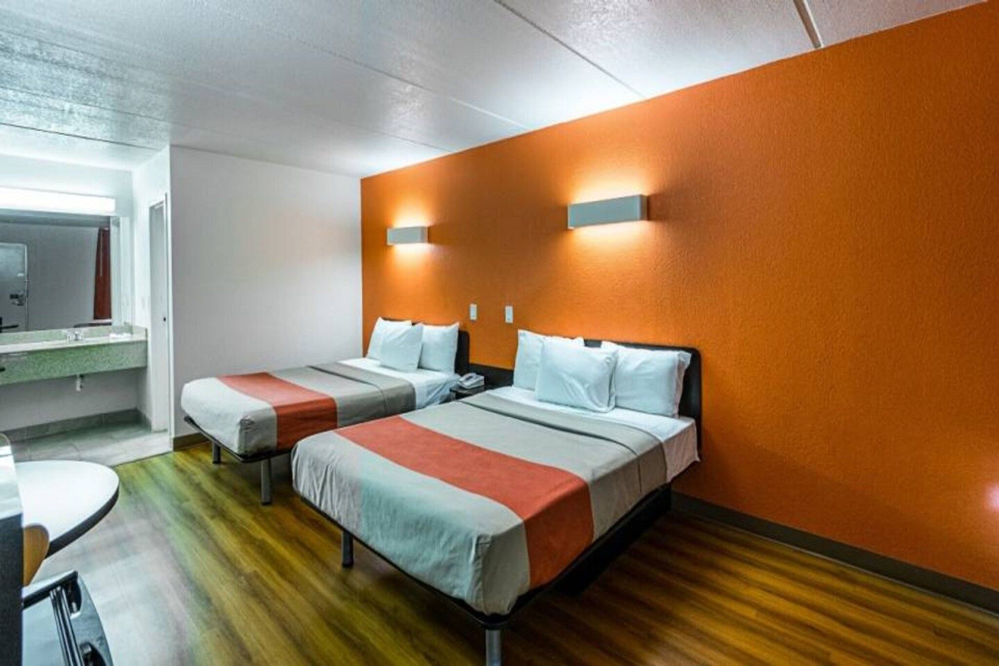 Quarto de hotel com duas camas, piso de madeira, banheiro com espelho e uma mesa pequena.