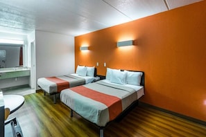 Quarto de hotel com duas camas, piso de madeira, banheiro com espelho e uma mesa pequena.