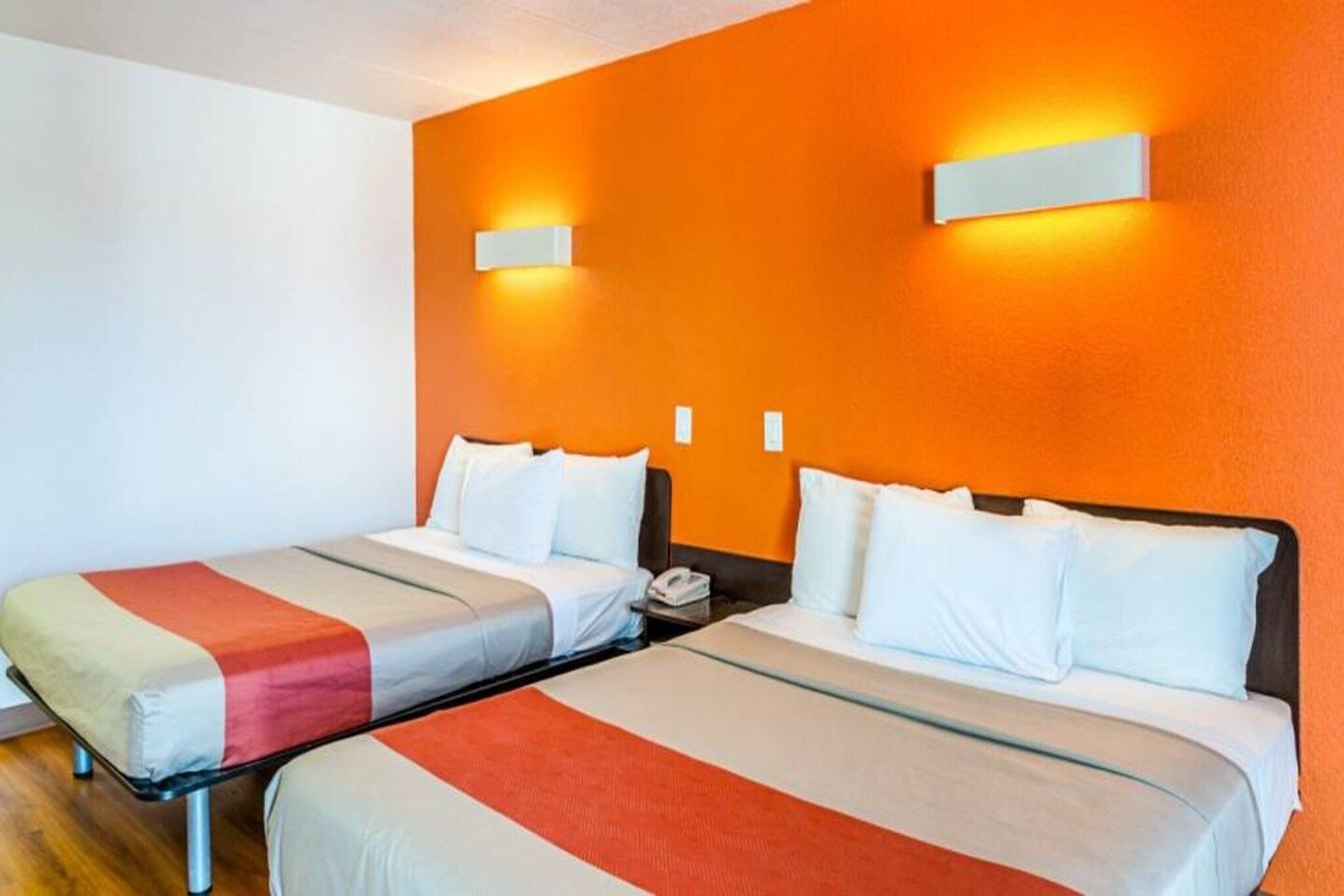A hotel room with two beds, each with a striped bedspread and white pillows. There are two wall-mounted light fixtures on the orange wall.
