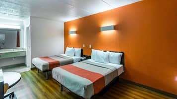 Quarto de hotel com duas camas, piso de madeira, banheiro com espelho e uma mesa pequena.