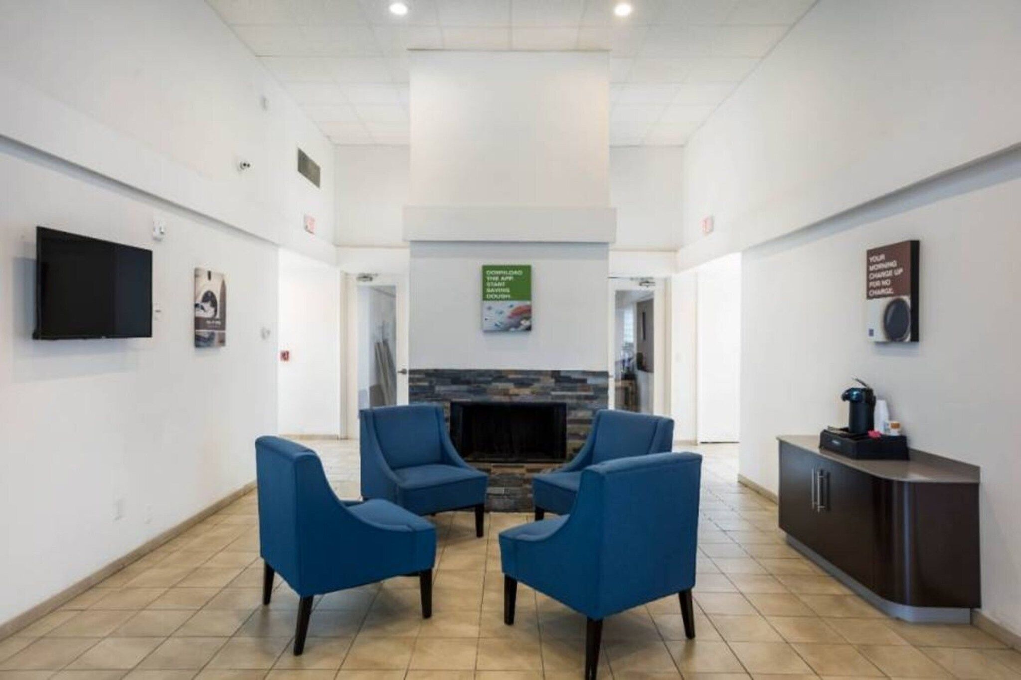 A lobby with a fireplace, blue chairs, a TV, and a coffee machine.