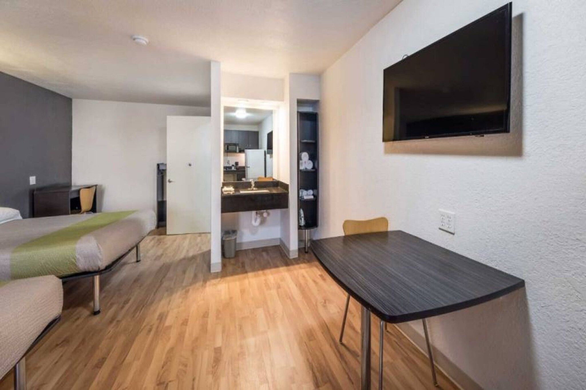 A compact hotel room with a bed, a small table, a TV mounted on the wall, and a kitchenette in the background.
