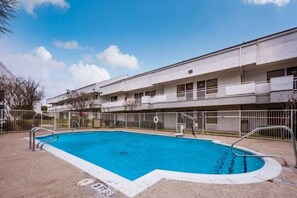 A rectangular swimming pool with a white border, surrounded by a metal fence, and a multi-story building with balconies in the background. - 2 FULL BEDS | SMOKING, KITCHEN (Irving TX)