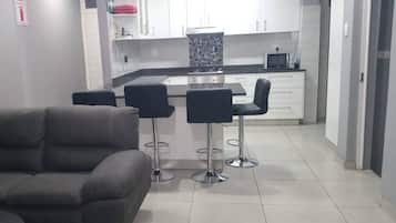 A modern kitchen with white cabinets, a black countertop, and a microwave. There are two black bar stools and a grey sofa in the living area.