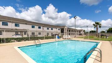 Piscina com água azul cristalina, rodeada por uma cerca, e um edifício com várias janelas ao fundo.