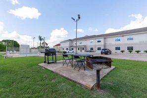 Un patio con una parrilla, sillas y una mesa, junto a un edificio con piscina.