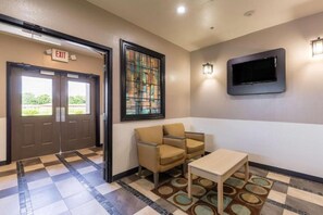 Un vestíbulo con suelo a cuadros, una zona de estar con dos sillones y una mesa, un televisor montado en la pared y una señal de salida.