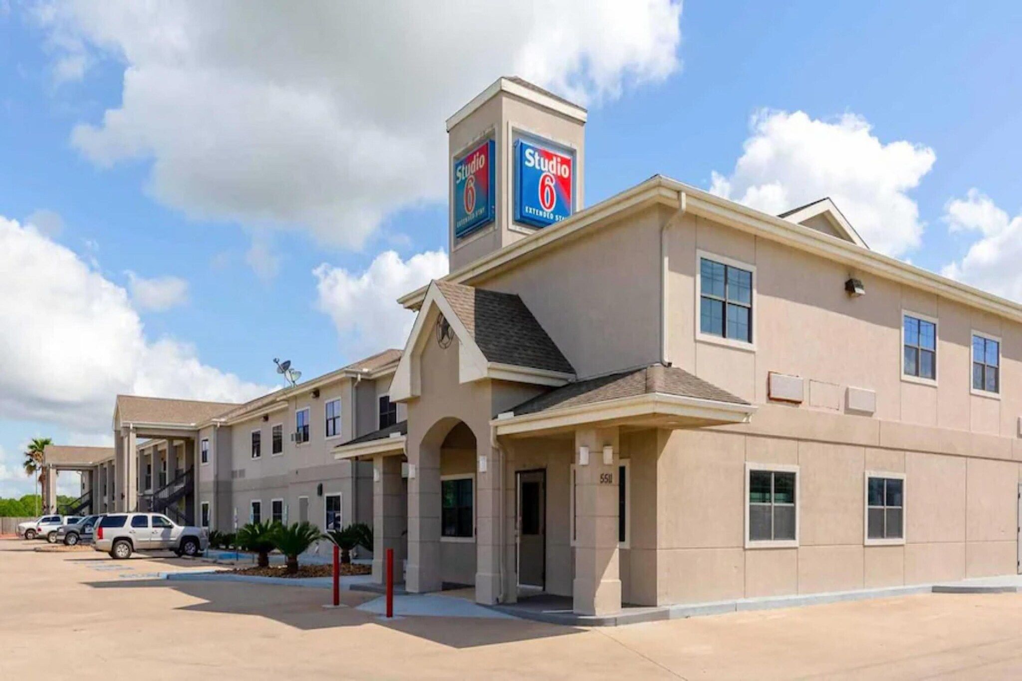 Un hotel de varios pisos con un llamativo letrero de Studio 6, un acceso cubierto y un estacionamiento con varios vehículos.