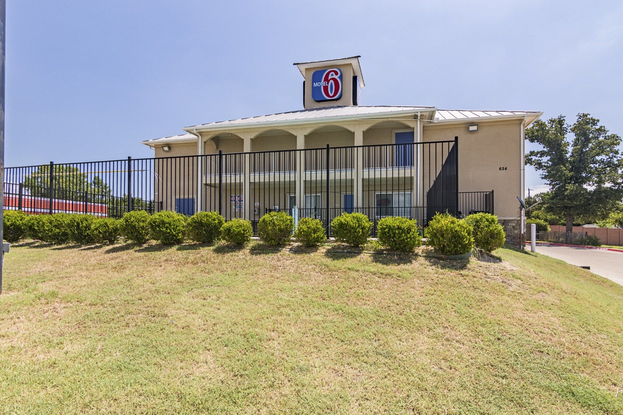 A Motel 6 building with a flat roof, a fenced parking area, and a well-maintained lawn.