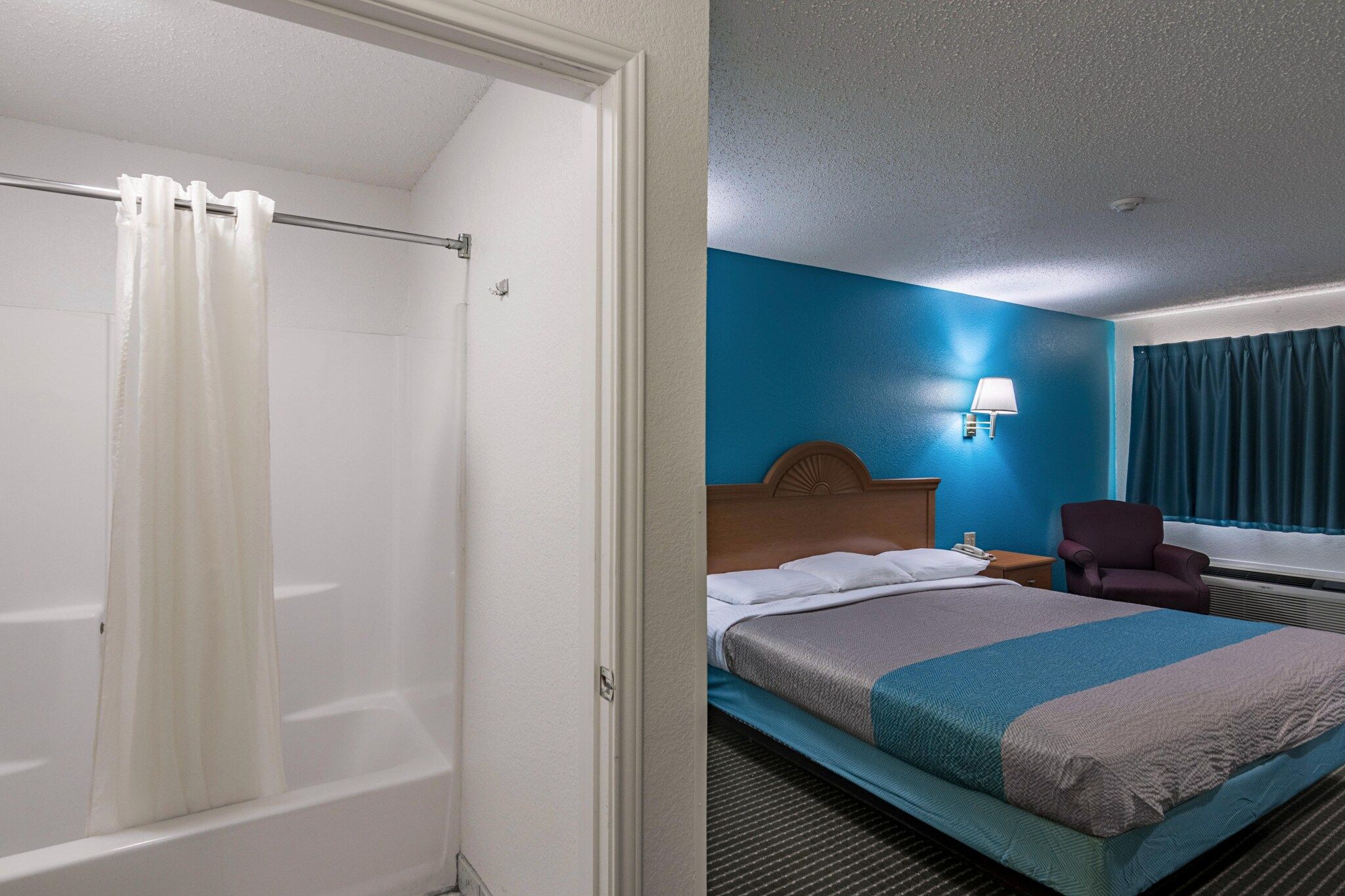 Ein Hotelzimmer mit Dusche, einem Bett mit Kopfteil, einem Nachttisch, einem Sessel und einem Fenster mit Vorhängen.