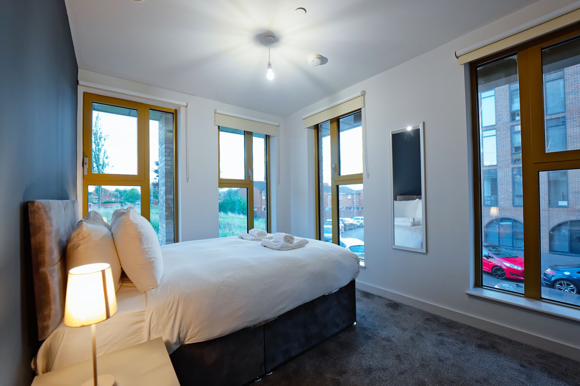A bedroom with a bed, a bedside lamp, a flat-screen TV, and large windows.