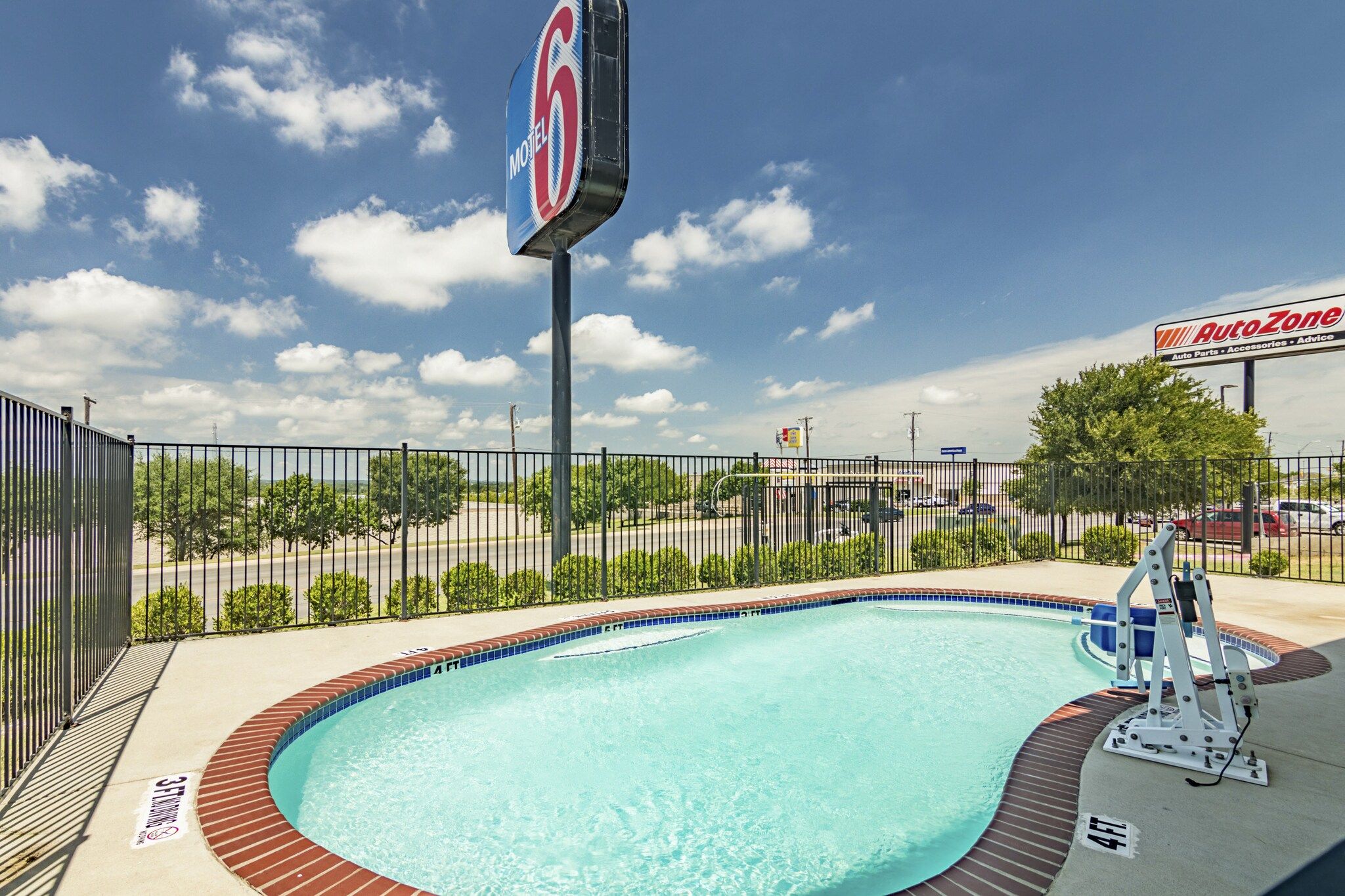 A swimming pool with a fence, a sign for Motel 6, and an AutoZone store in the background.