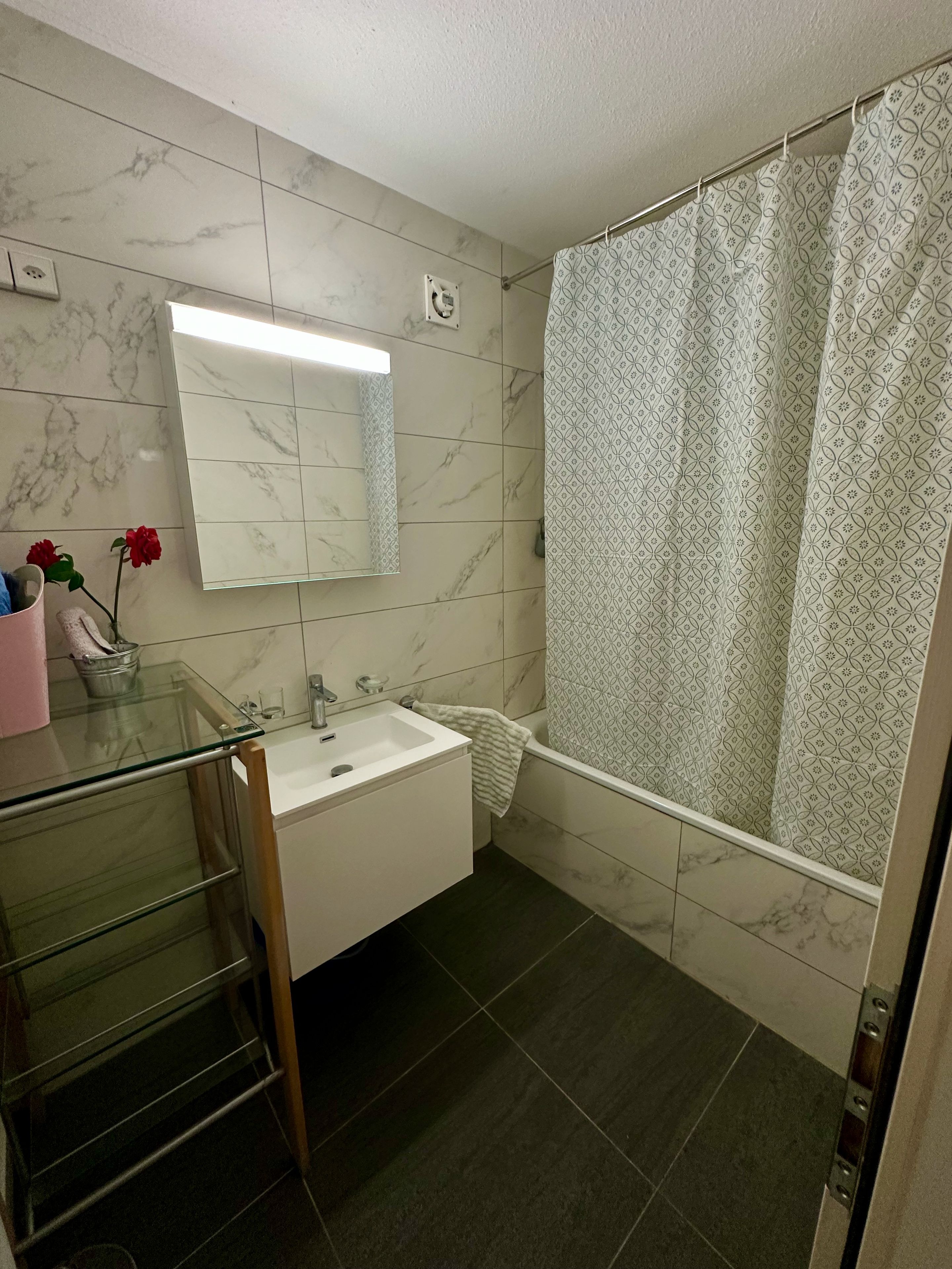 Een badkamer met een wastafel met glazen blad, een spiegel, een douchegordijn en een betegelde vloer.