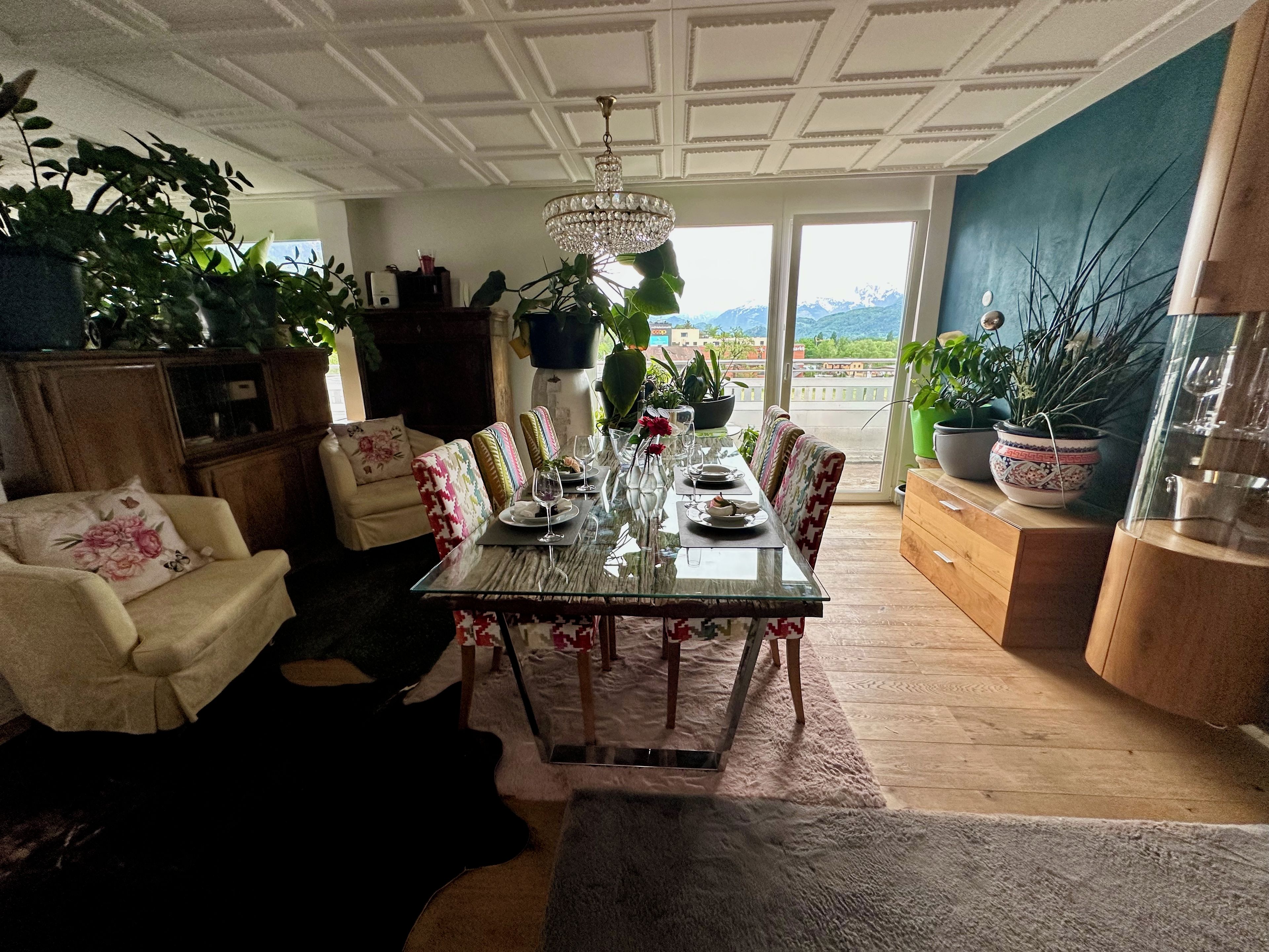 A dining area with a glass table, chairs, and a chandelier. There are potted plants and a view of the outdoors.