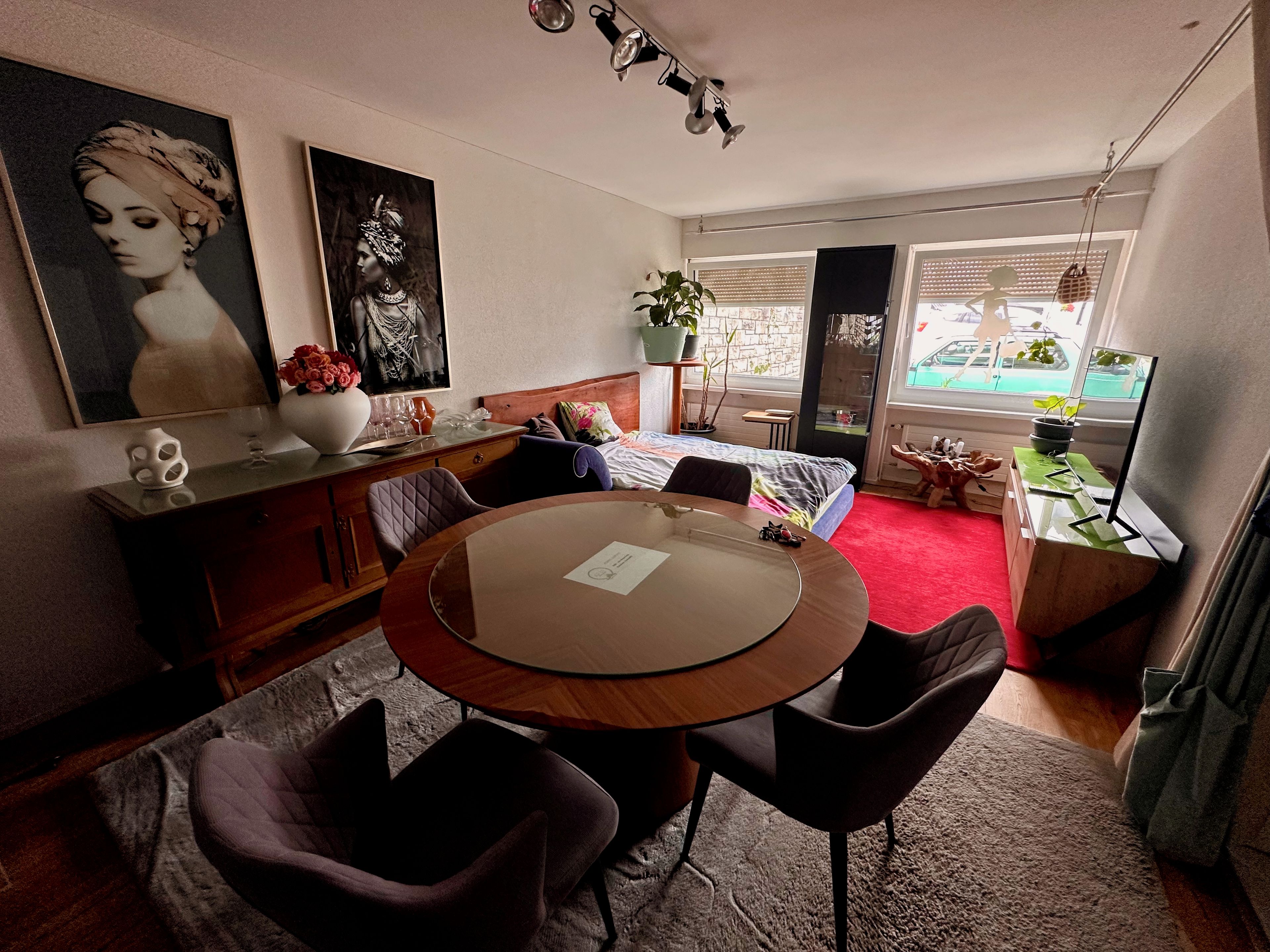 A room with a round wooden table, two chairs, a bed with a colorful bedspread, a small side table with a plant, and a window with a view of the outside.