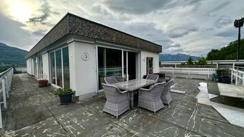 A rooftop terrace with a dining table and chairs, a small building with a glass door, and a mountainous backdrop.