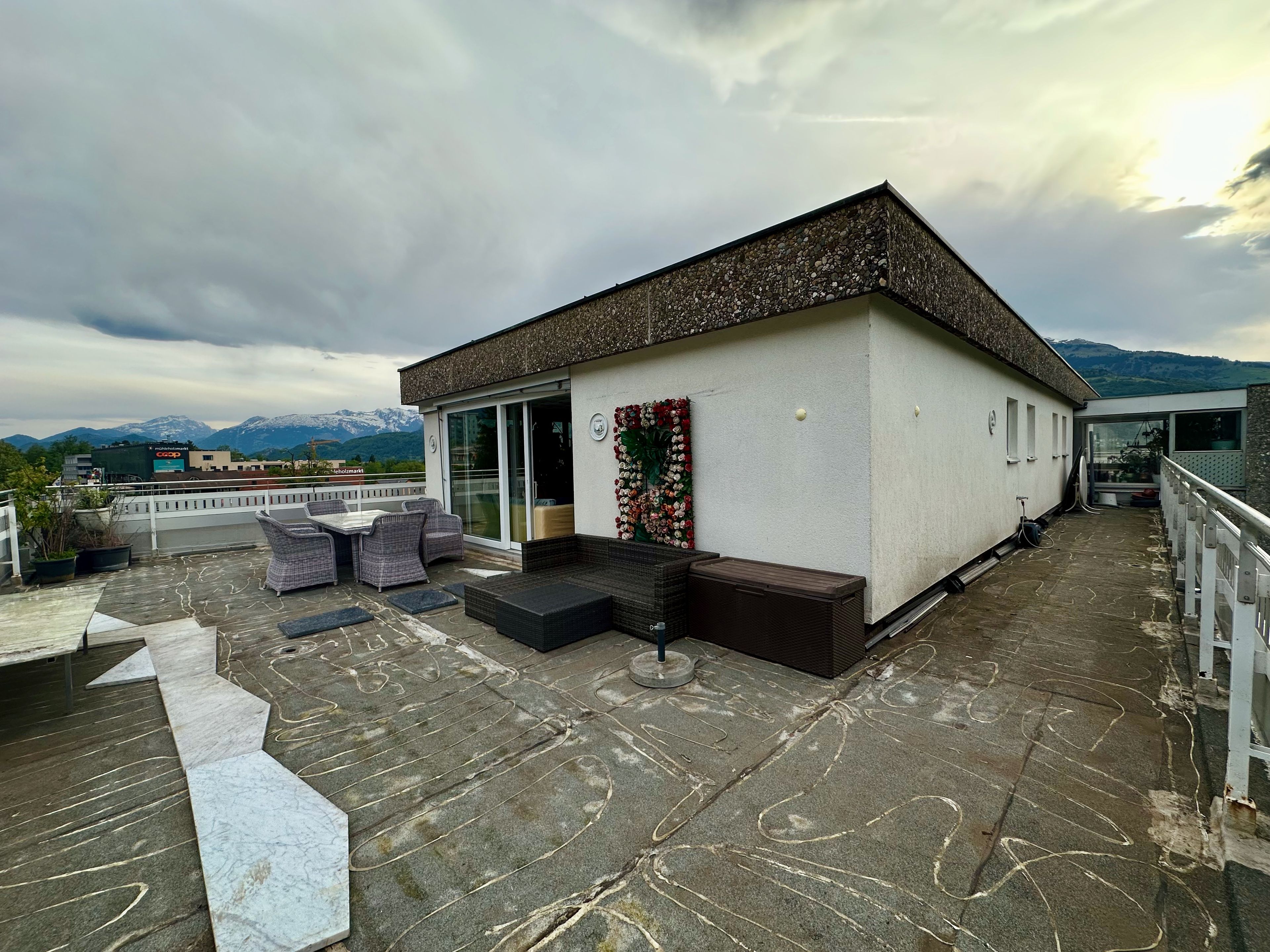 Una terraza en la azotea con mobiliario exterior, un edificio pequeño con puerta de cristal y vistas a la montaña.