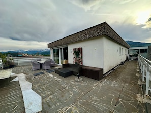 Una terraza en la azotea con mobiliario exterior, un edificio pequeño con puerta de cristal y vistas a la montaña.