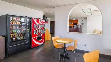 Un distributeur automatique de collations et un autre de boissons, notamment de Coca-Cola, une petite table avec des chaises et un miroir accroché au mur.