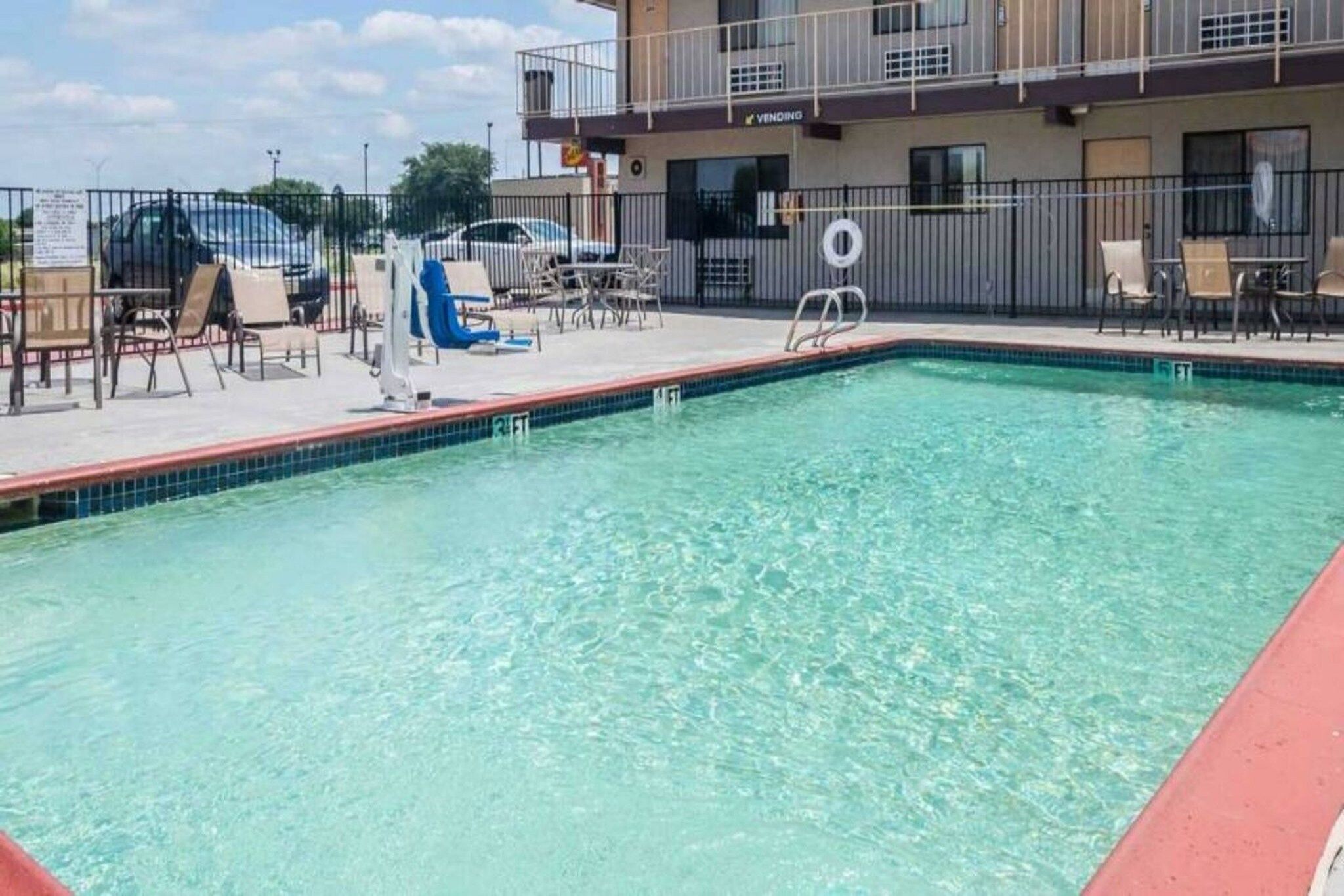 A swimming pool with a tiled edge, lounge chairs, and a fenced area with a building in the background.