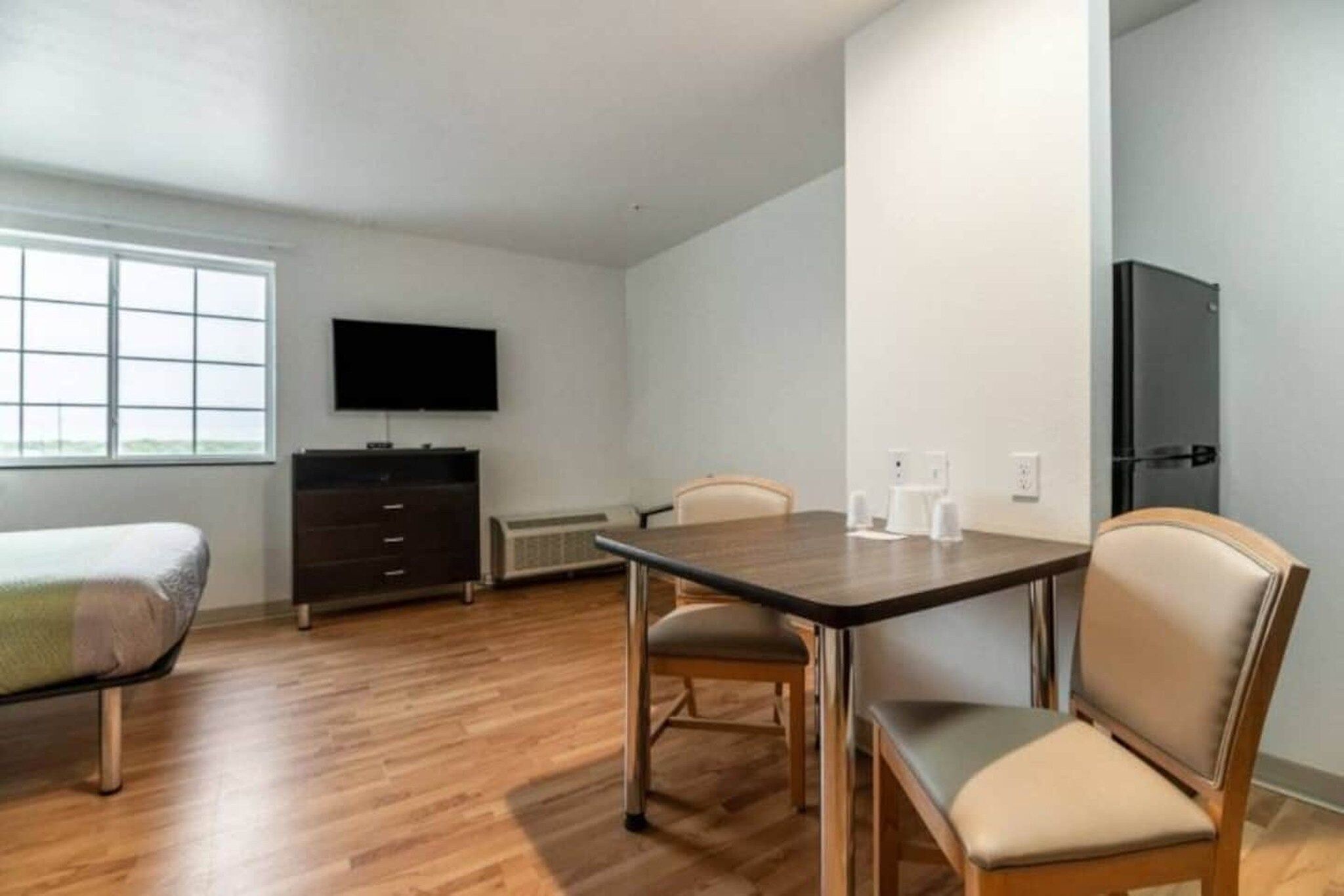 A room with a wooden floor, a TV mounted on the wall, a bed, a table with chairs, and a wardrobe.