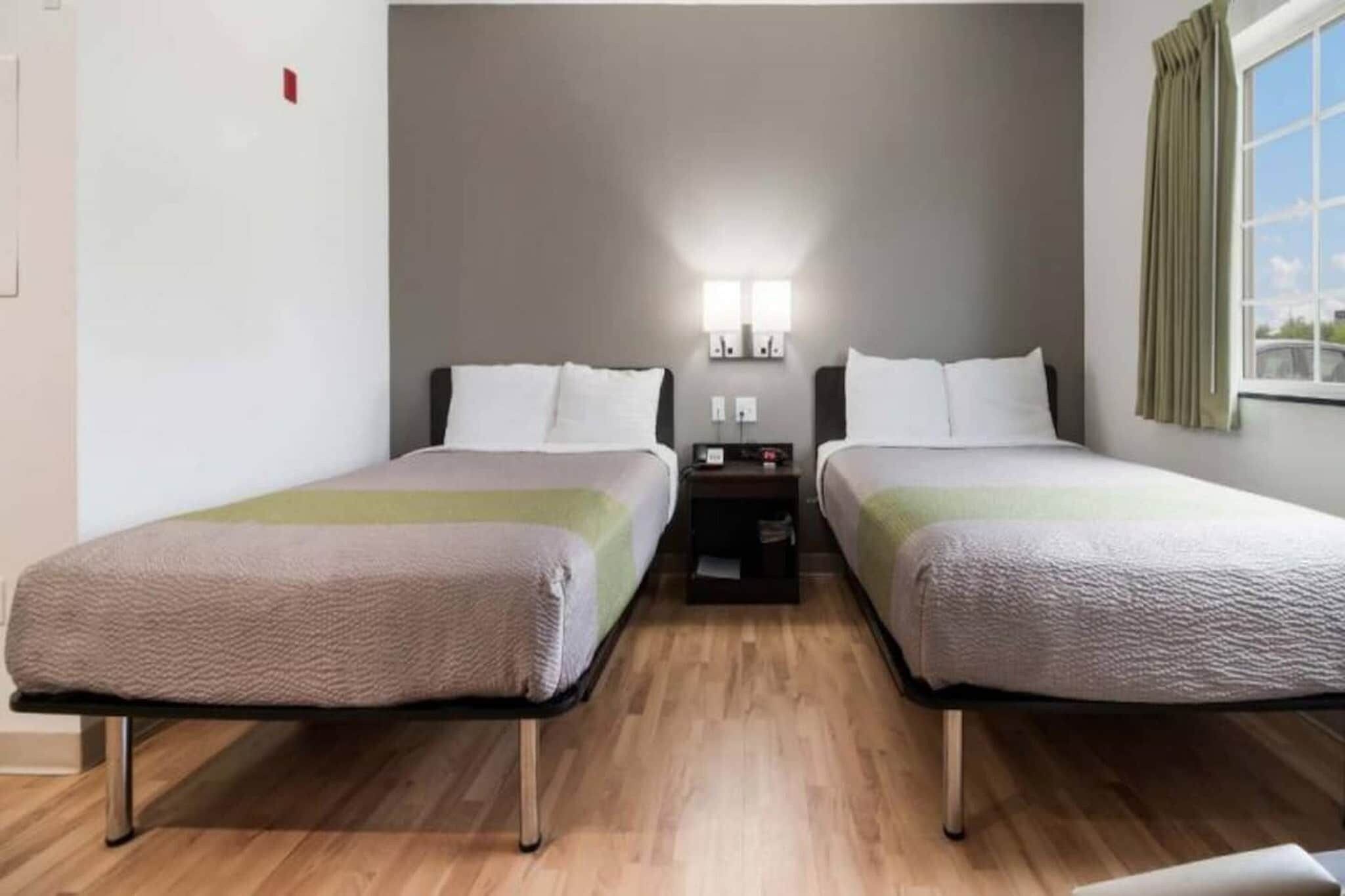 A hotel room with two beds, a nightstand, a lamp, and a window with curtains.