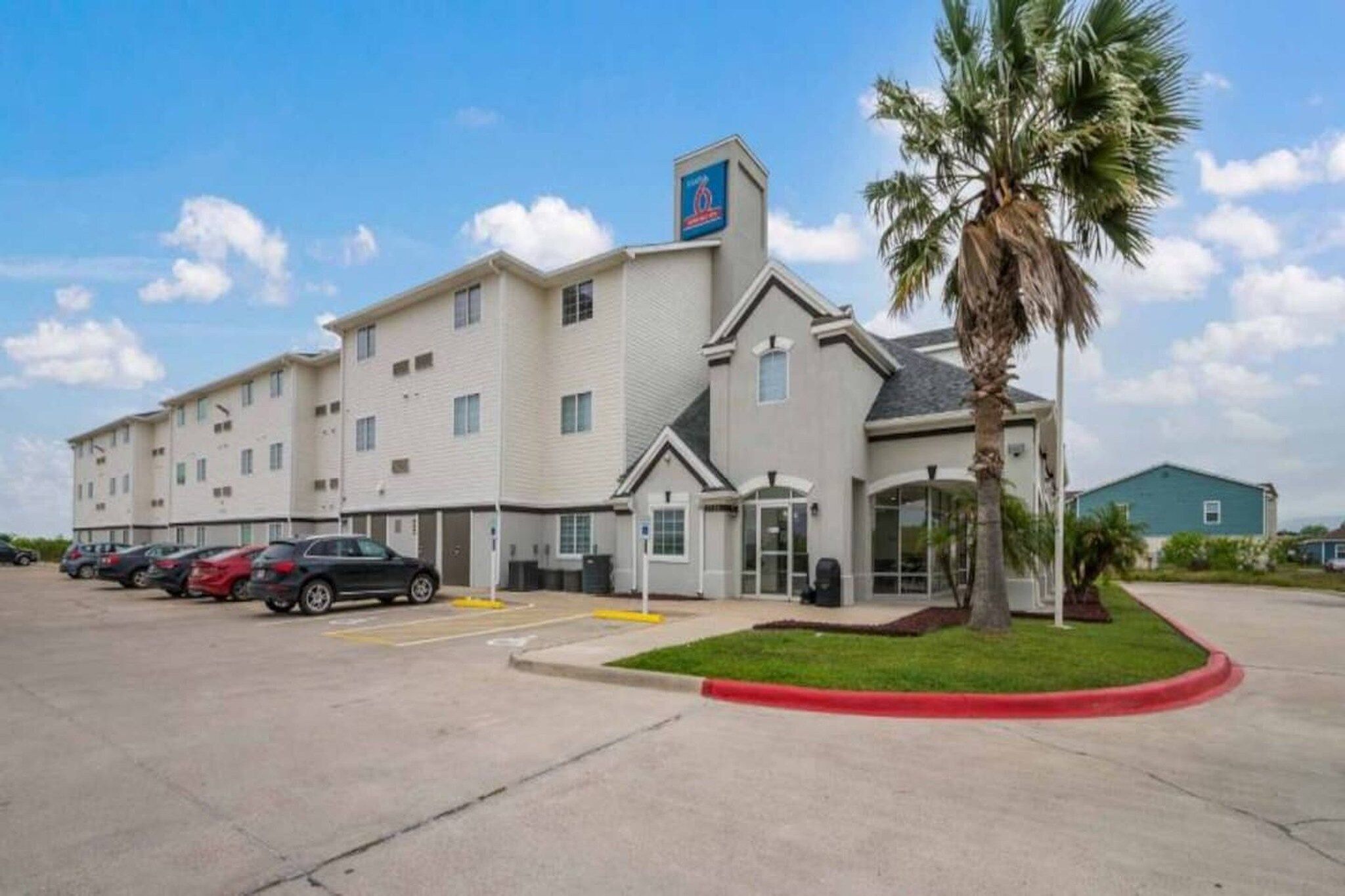 A hotel with a parking lot, a palm tree, and a clear sky.