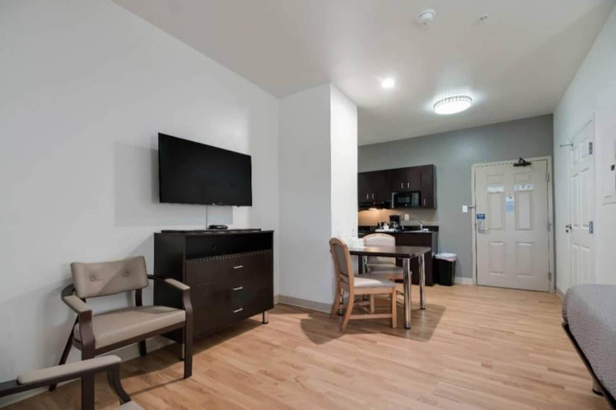 A compact living space with a flat-screen TV mounted on the wall, a dining area with a table and chairs, and a kitchenette in the background.