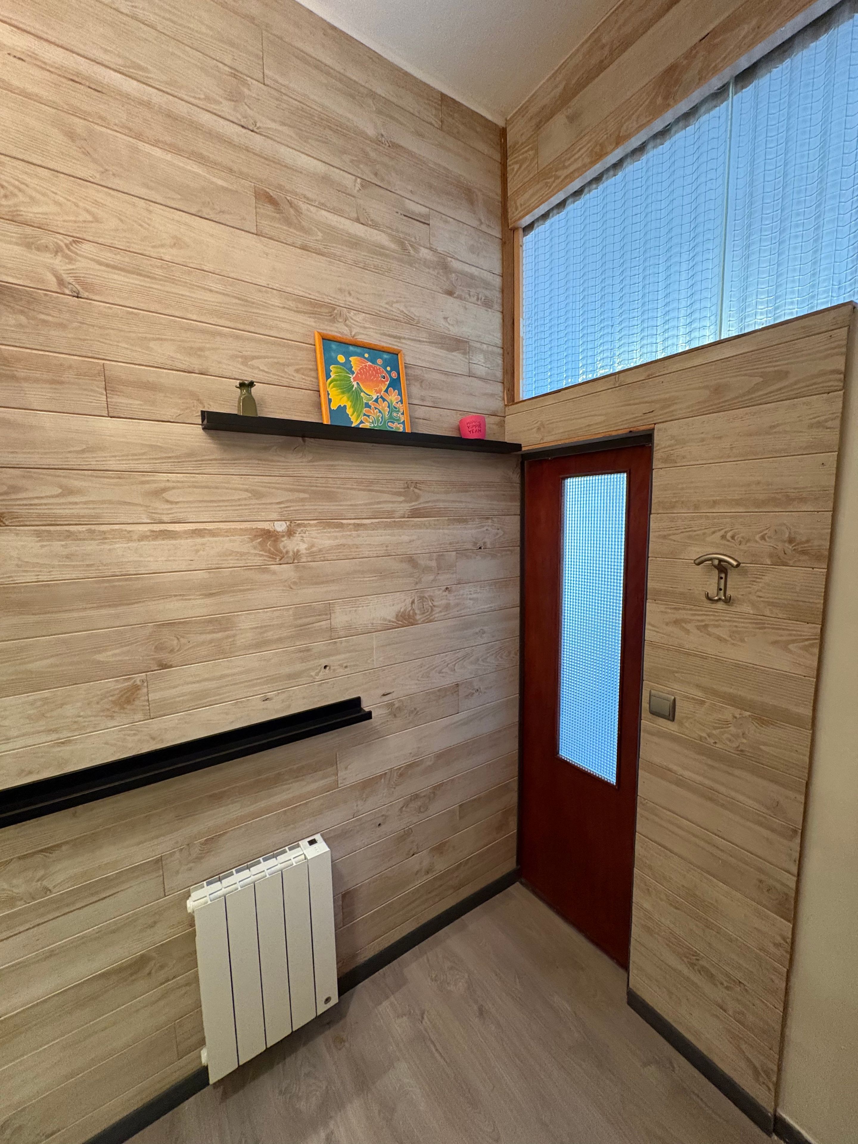 Een gang met houten paneelwanden, een radiator, een deur met raam en een plank met een ingelijst beeld.