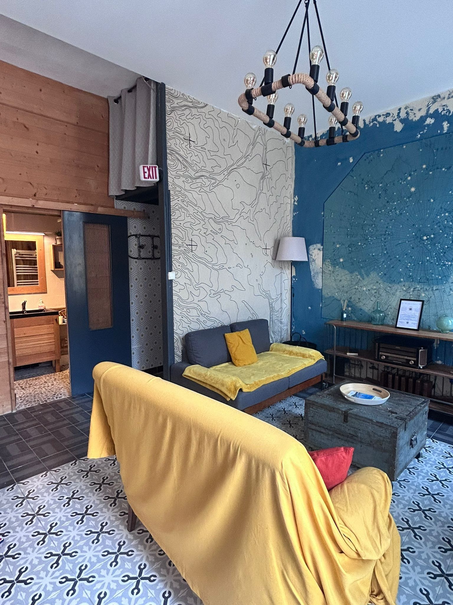 Een moderne woonkamer met een bank, een salontafel en een kroonluchter. Er hangt een kaart aan de muur en een deur met een nooduitgangsbordje.
