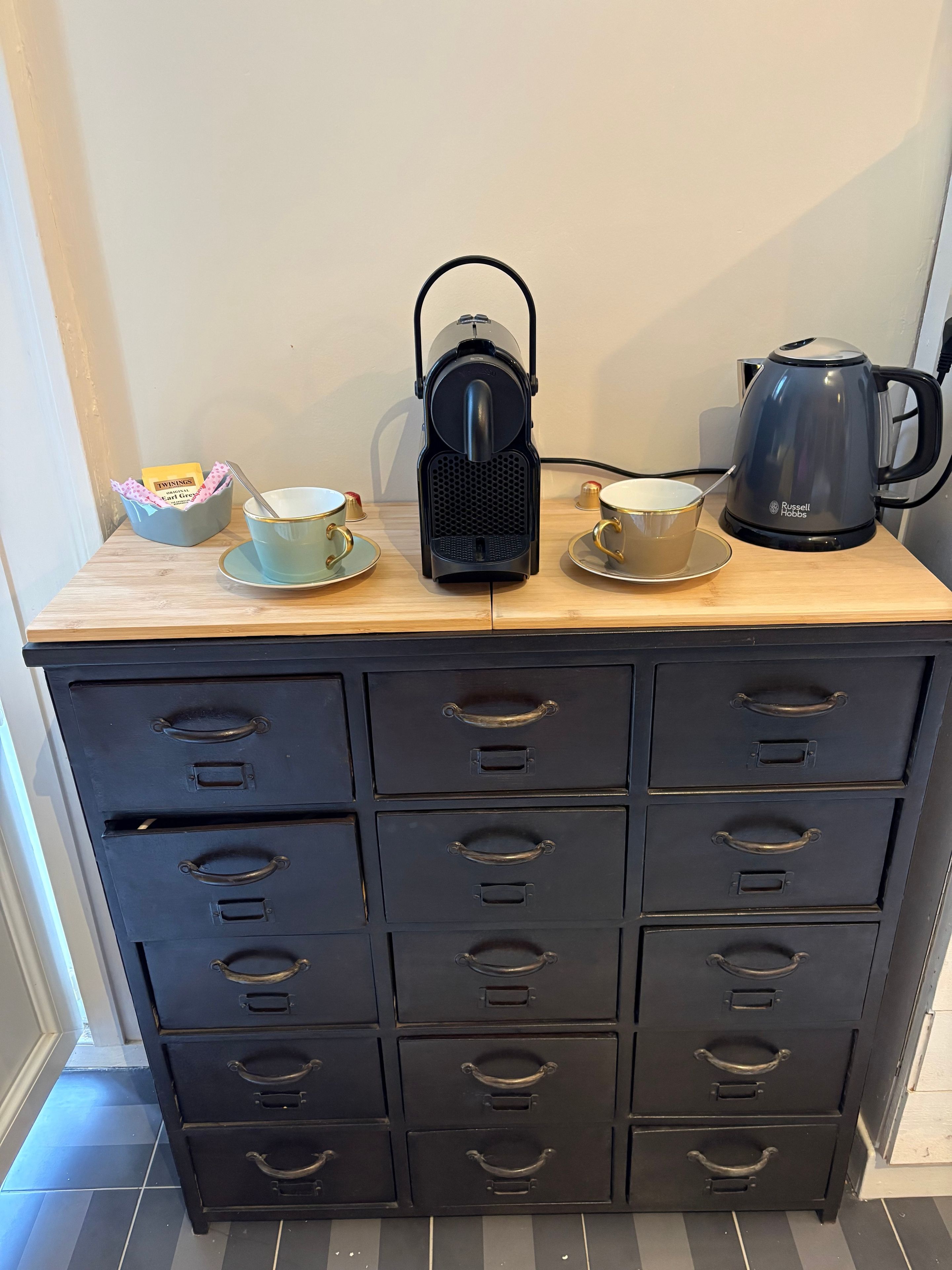 Een kitchenette met een koffiezetapparaat, twee kopjes en een kom op een houten aanrecht.