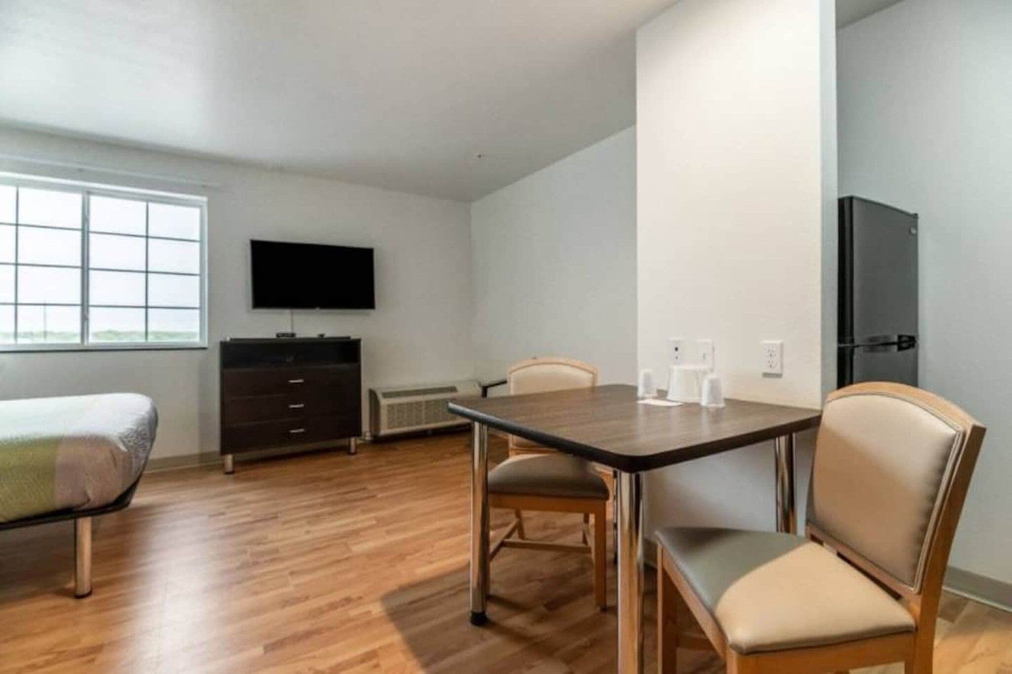 A room with a wooden floor, a TV mounted on the wall, a bed, a table with chairs, and a wardrobe.