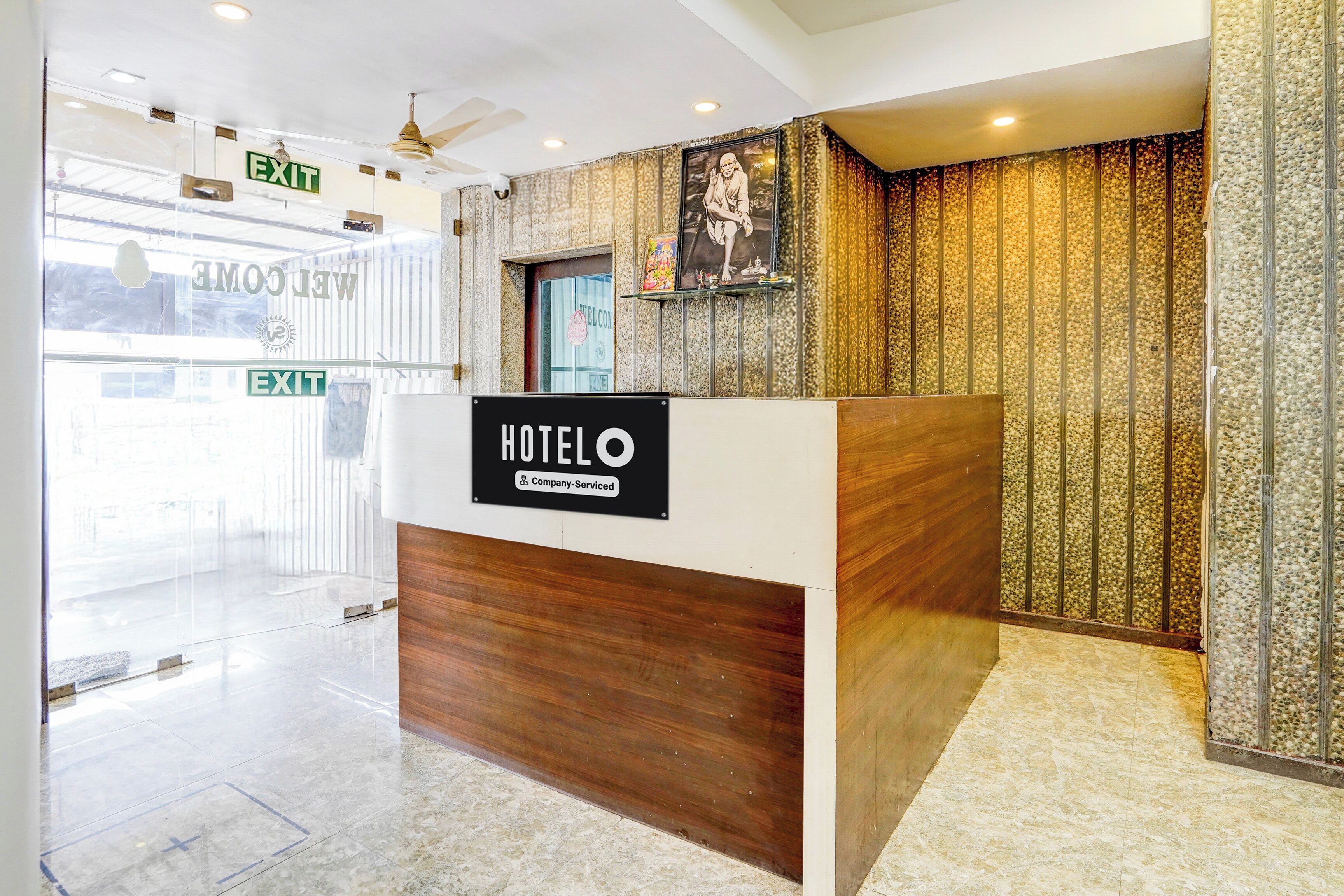 Mostrador de recepción de hotel con un cartel que indica servicio de empresa. Se ven dos salidas al fondo, una con la señalización 'EXIT' y otra con un cartel de 'BIENVENIDA'.