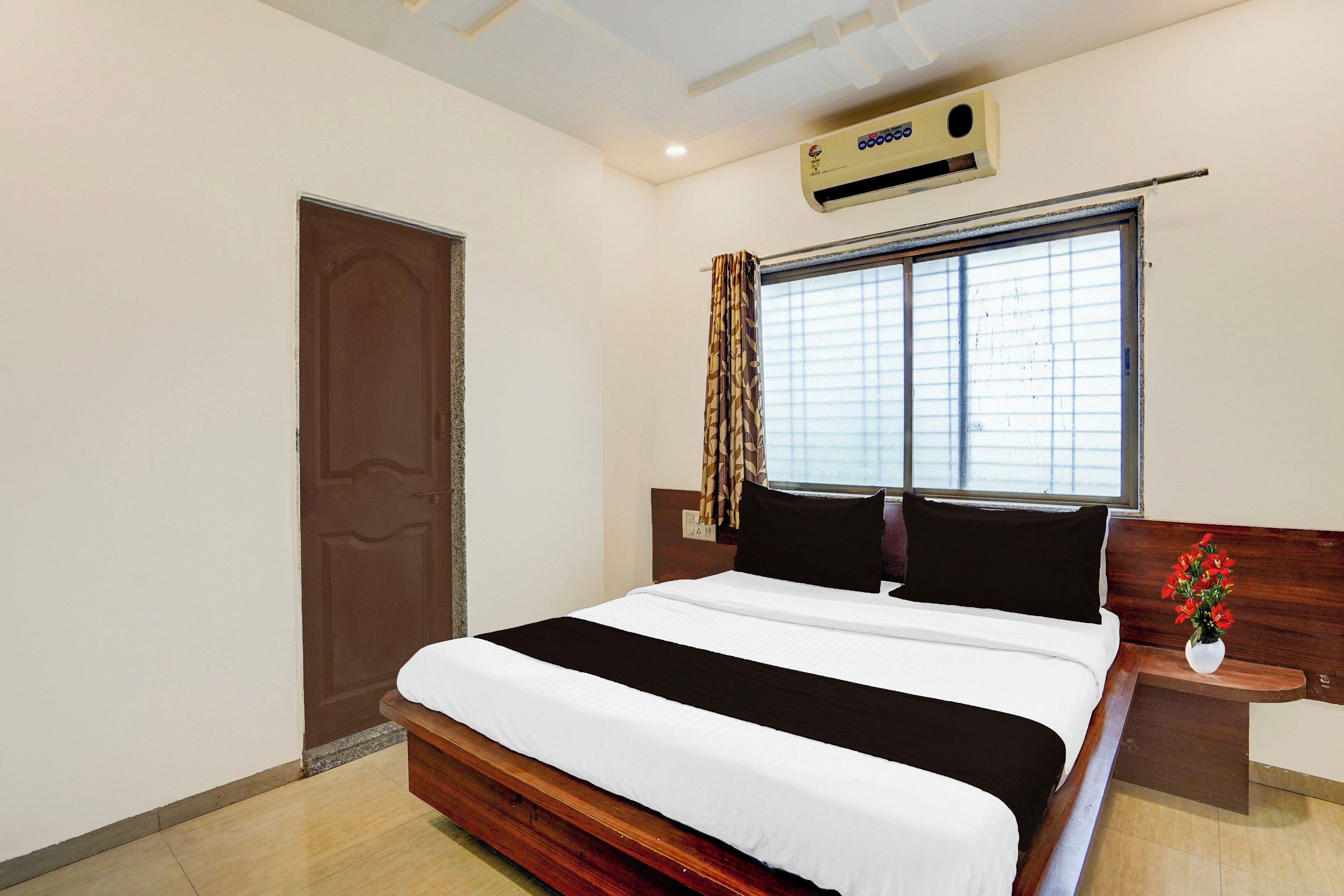 A hotel room with a bed, a window with curtains, a wall-mounted air conditioner, and a wooden headboard.