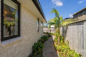 A narrow concrete pathway flanked by a brick wall and a wooden fence, with lush green plants and a palm tree. - Waterways No. 6 - Whitianga (Whitianga)