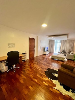 A spacious living room with a wooden floor, a black office chair, a sofa, and a television. - Finewood Residency View Palace Apartment (Nairobi)