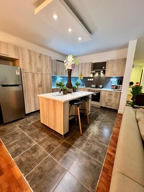 A modern kitchen with a central island, wooden cabinets, and a tiled backsplash. - Finewood Residency View Palace Apartment (Nairobi)
