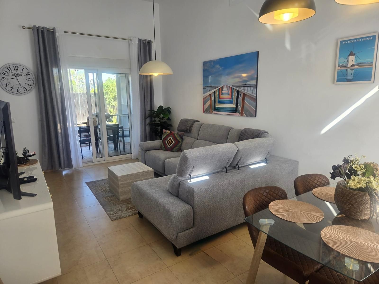 A modern living room with a grey sofa, a glass dining table with placemats, a television, and a painting of a bridge.