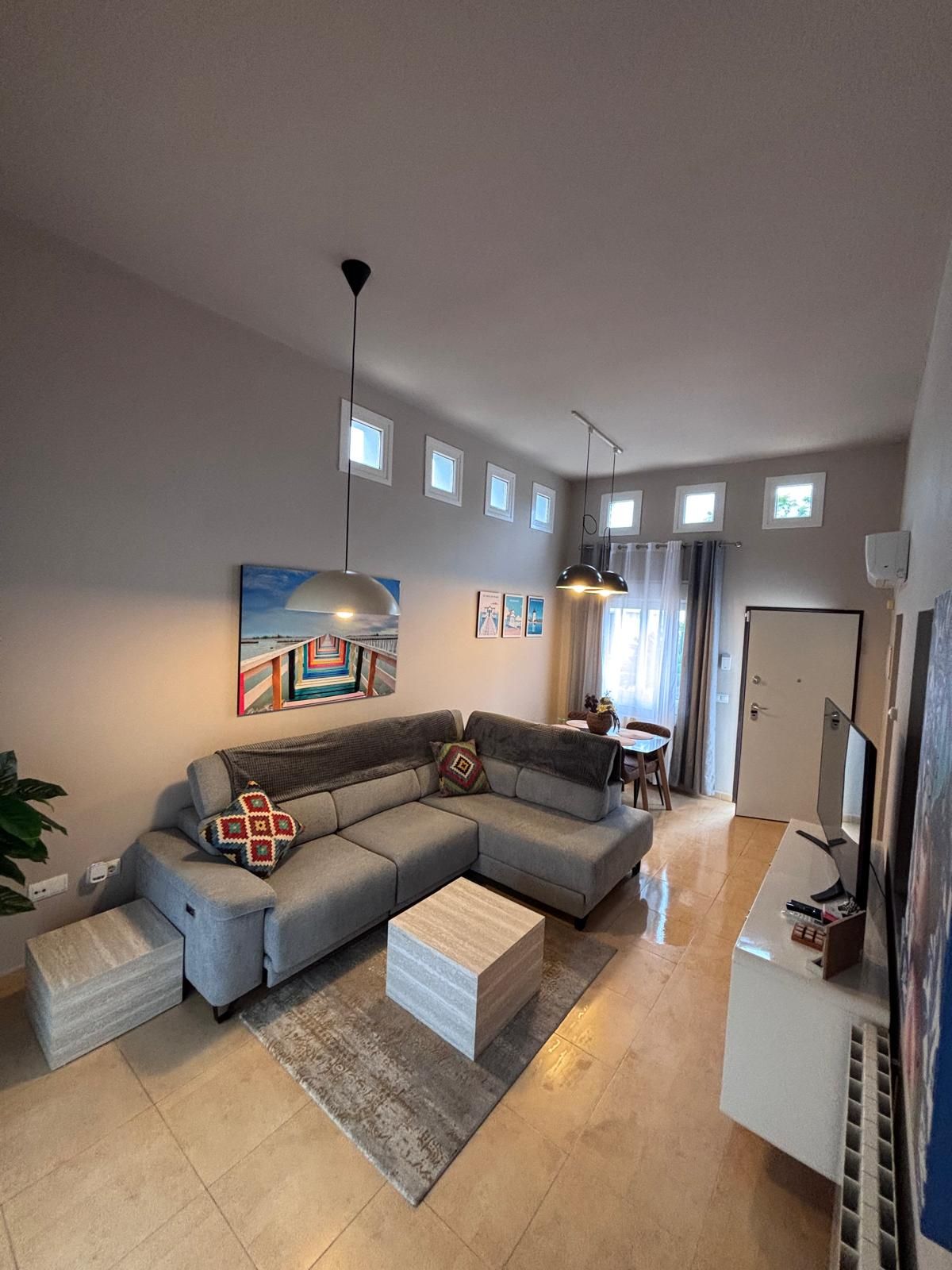 Un salon moderne avec un canapé d’angle, une table basse et un téléviseur à écran plat. De nombreuses fenêtres laissent entrer la lumière naturelle et offrent une vue sur l’extérieur.