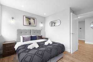 A modern bedroom with a bed, nightstands, a headboard, a wall-mounted mirror, and a door leading to another room. - The Isleworth 2BR 2BA with Gated Parking & Balcony (Isleworth)