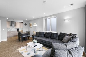 A modern living room with a grey sofa, a dining table with chairs, a kitchen in the background, and a large window. - The Isleworth 2BR 2BA with Gated Parking & Balcony (Isleworth)