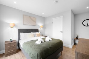 A neatly made bed with a green blanket, two bedside lamps, a small bedside table with a vase, and a wall-mounted mirror. - The Isleworth 2BR 2BA with Gated Parking & Balcony (Isleworth)