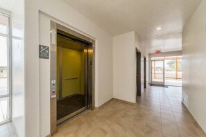 Un couloir intérieur avec un ascenseur à l’arrêt, une porte portant un panneau indiquant la sortie de secours, et un sol carrelé.