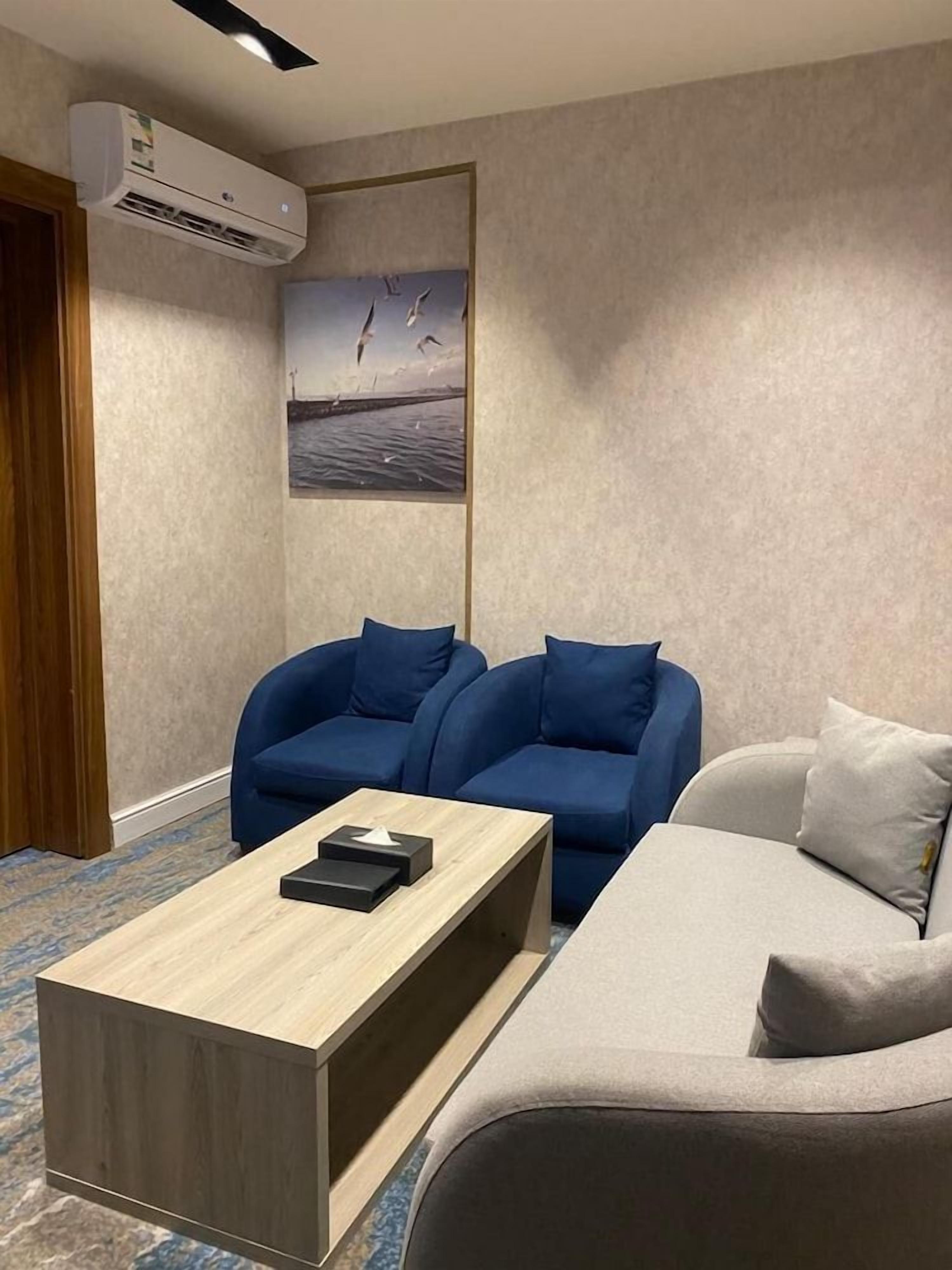 Una habitación de hotel con una mesa de centro de madera, dos sillones azules, un sofá gris, un cuadro de pájaros y un equipo de aire acondicionado.