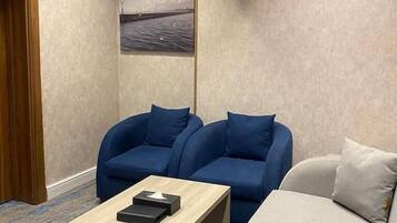 Una habitación de hotel con una mesa de centro de madera, dos sillones azules, un sofá gris, un cuadro de pájaros y un equipo de aire acondicionado.