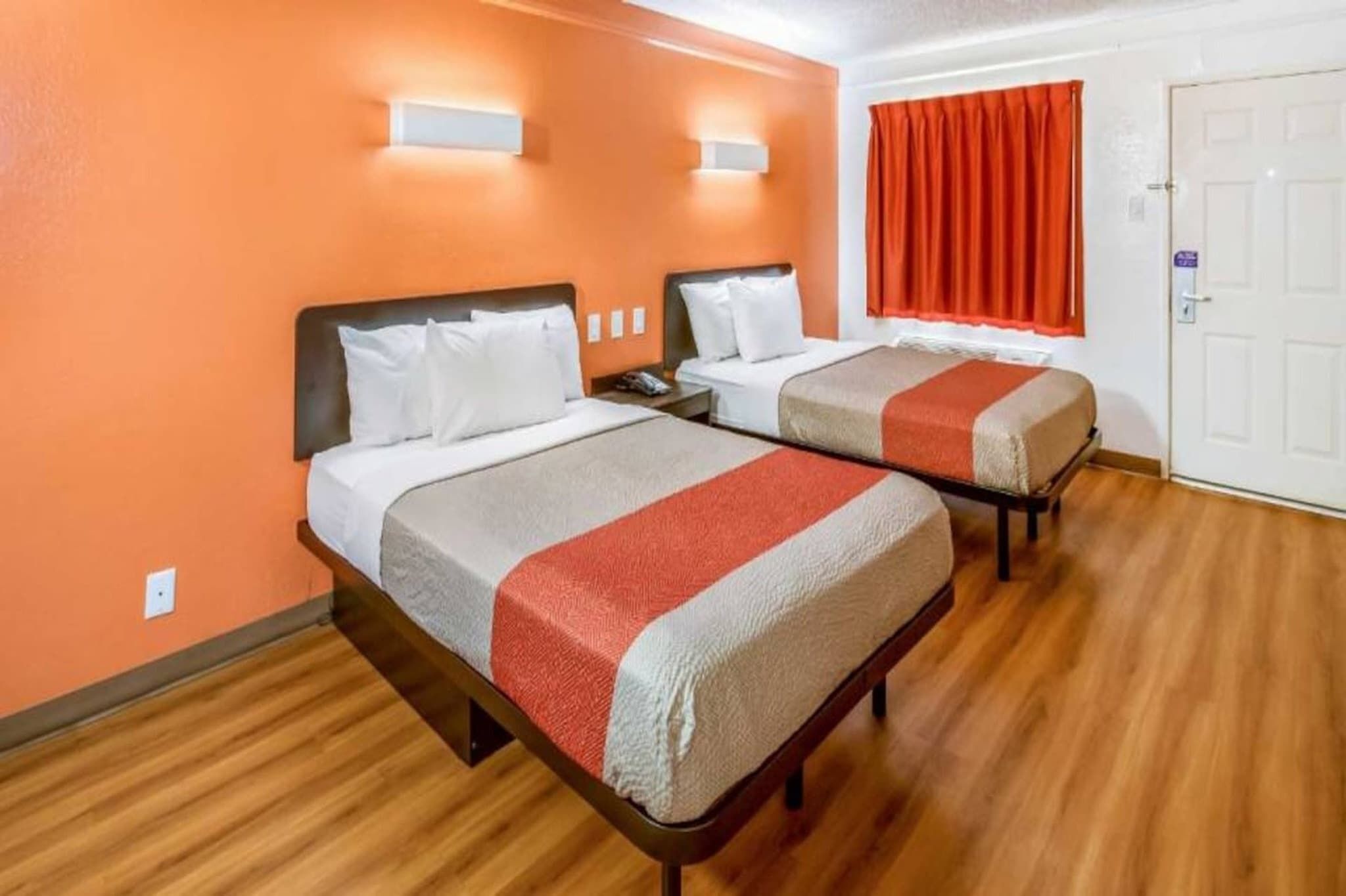A hotel room with two beds, wooden flooring, and a red curtain.