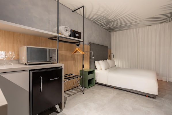 Habitación de hotel moderna con una cama, un escritorio con microondas, un mini refrigerador, una percha para ropa y una mesita de noche.