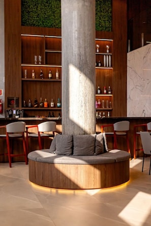 Un área de bar moderna con disposición circular de asientos, estantes de madera con botellas y un pilar grande.