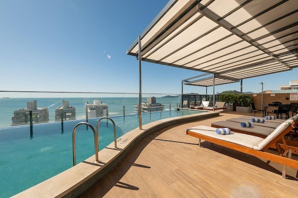 Piscina infinita no terraço com espreguiçadeiras e área de estar coberta, com vista para um corpo de água e edifícios.